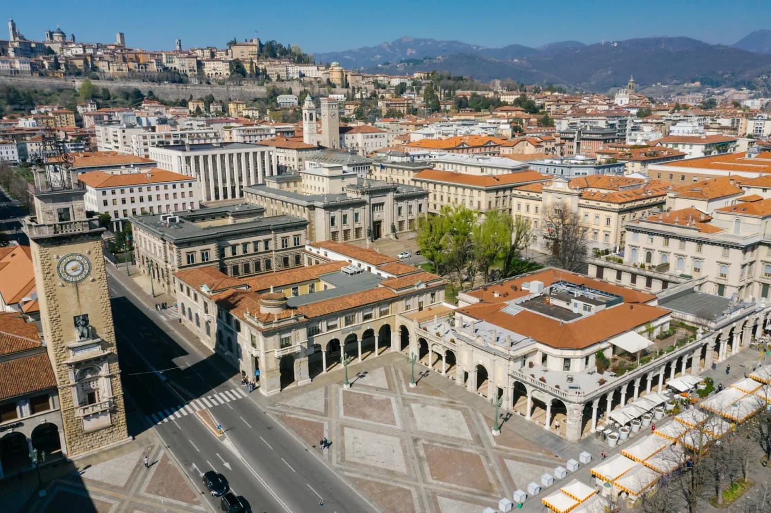 Luftaufnahme der Piazza Vecchia in Bergamo mit Uhrturm, Arkaden, historischen Gebäuden und rotdachigen Häusern vor Hügeln in Norditalien