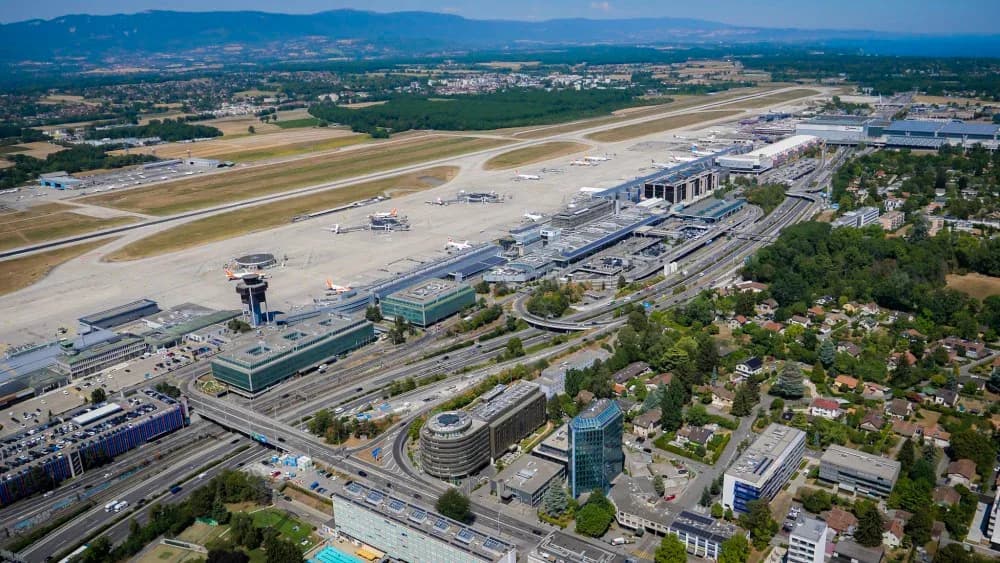 Luftaufnahme Flughafen Genf mit Terminals, Rollfeld, parkenden Flugzeugen, Kontrollturm und angrenzender Stadtlandschaft