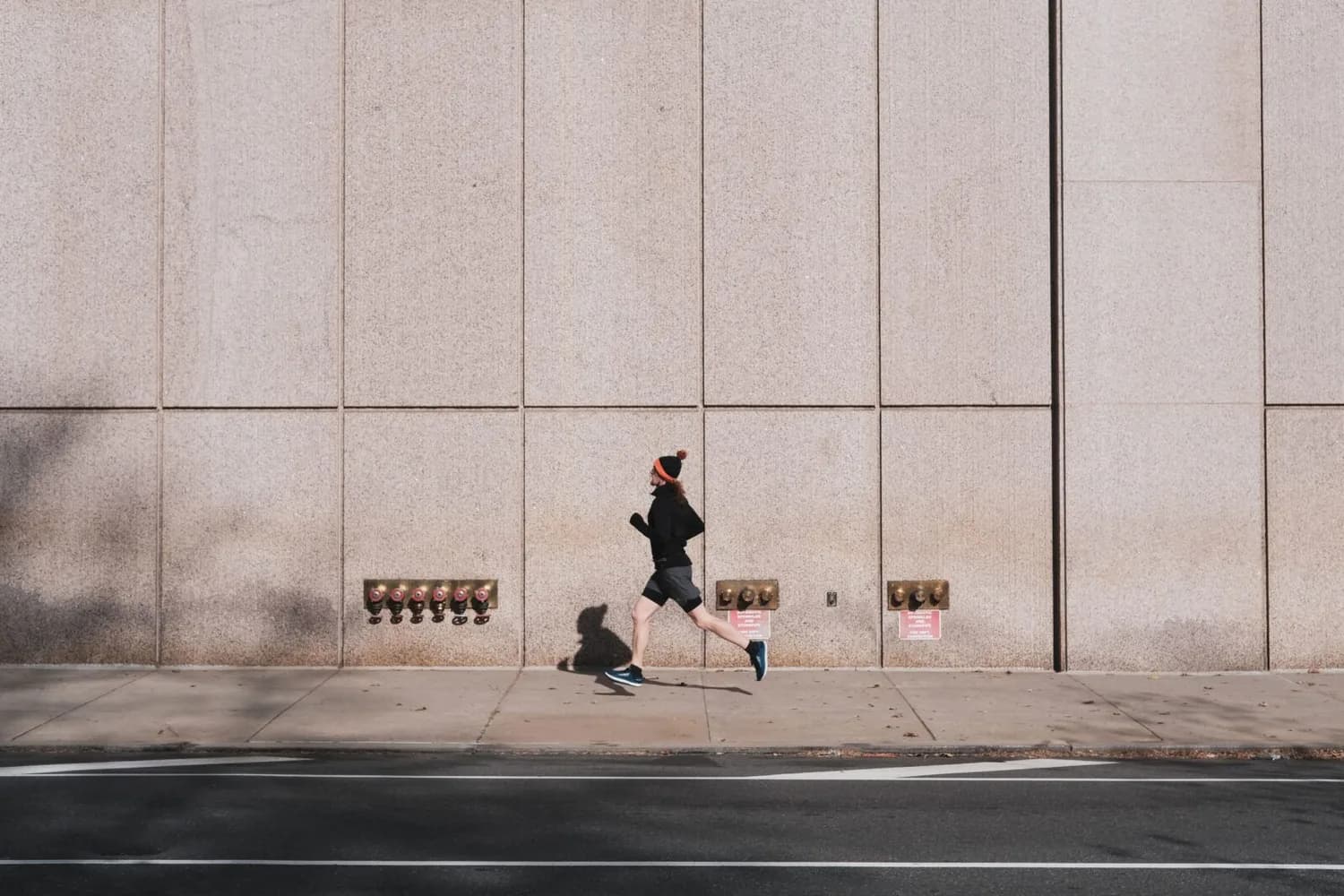 Jogger in Sportkleidung läuft vor großer Betonfassade am Flughafen MUC, urbanes Straßenmotiv, Seitenansicht, Bewegung und Schattenwurf