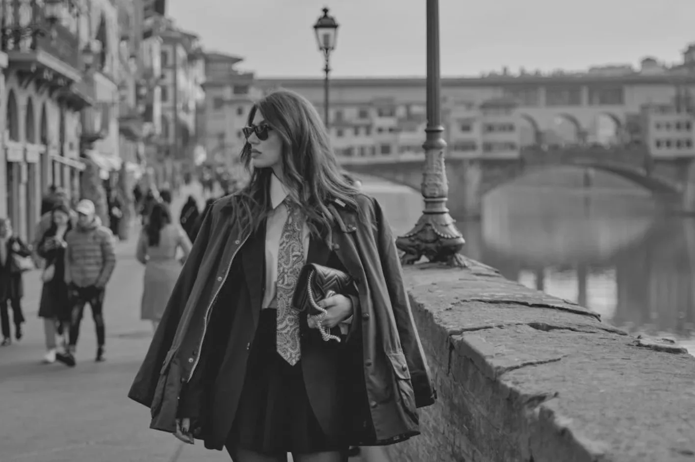 Frau mit Sonnenbrille und Handtasche spaziert an der Uferpromenade in Florenz mit Blick auf die Ponte Vecchio, Modeoutfit, Toskana