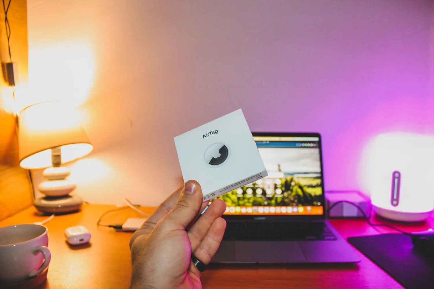 Hand hält Verpackung eines Apple AirTag vor Laptop auf Schreibtisch mit Lampe, Maus und Tasse