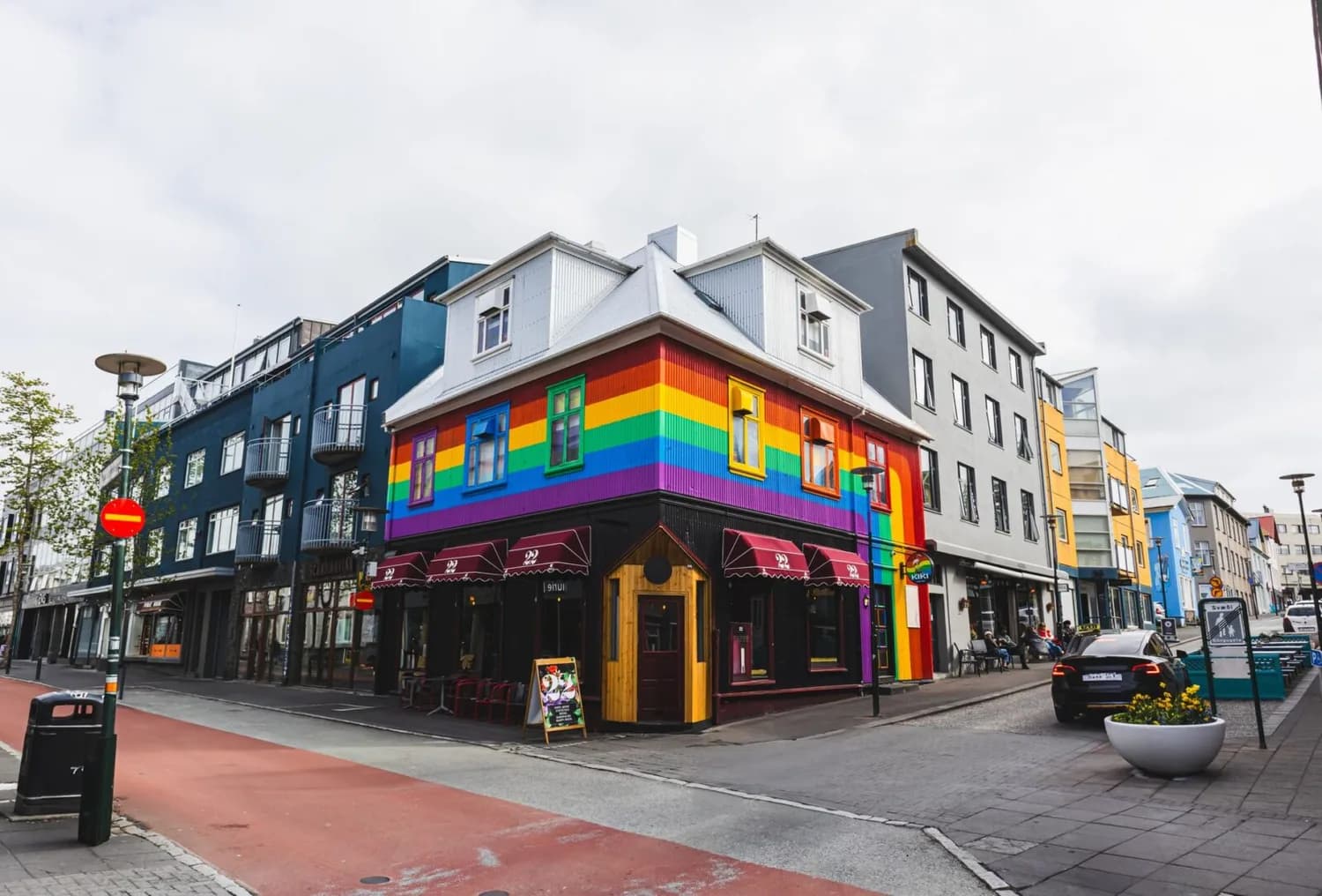 Bunt gestrichenes Eckgebäude mit Regenbogenstreifen und Café im belebten Straßenviertel von Reykjavík, Fußgängerbereich, umliegende moderne Häuser und parkender Wagen