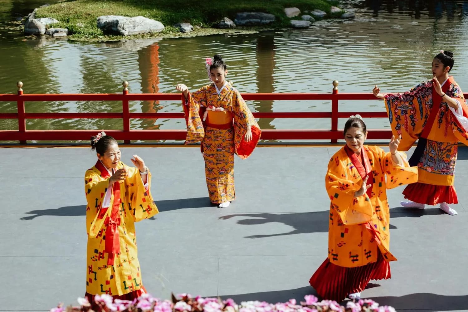 Vier Tänzerinnen in bunten japanischen Kimonos führen traditionelle Tanzbewegungen auf einer Bühne vor einem Teich mit roter Brüstung aus