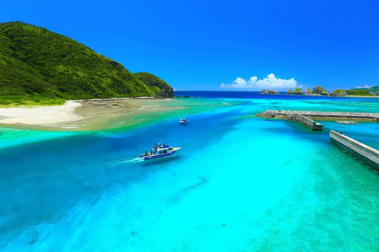 Türkisblaues Meer mit Bootsverkehr zum Hafen einer grünen Insel, Sandstrand und felsigen Inselriffen am Horizont (c) Okinawa Convention & Visitors Bureau
