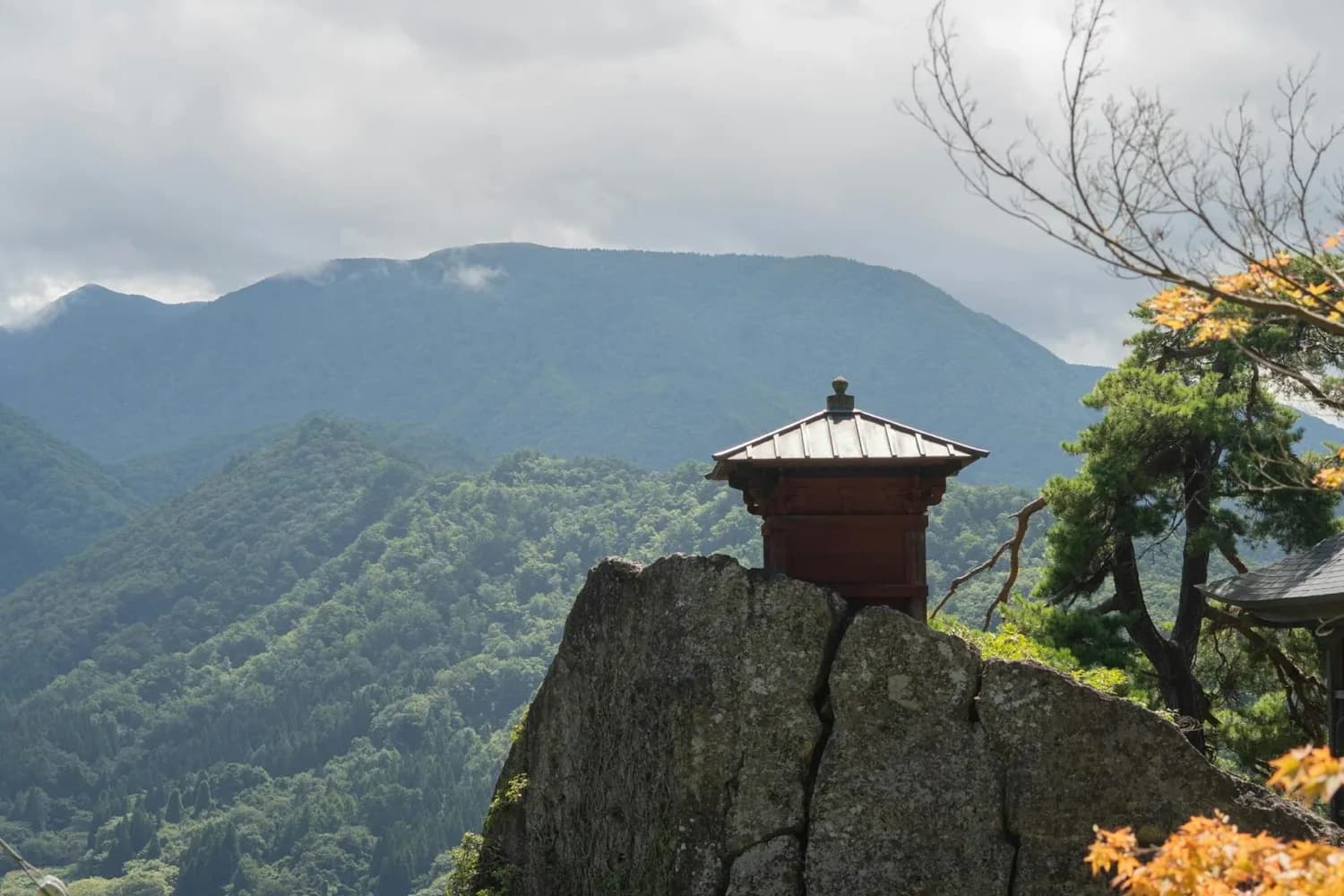 Bergtempel auf Felsvorsprung in Yamadera, Japan, mit grün bewaldeten Bergen und Nebel im Hintergrund