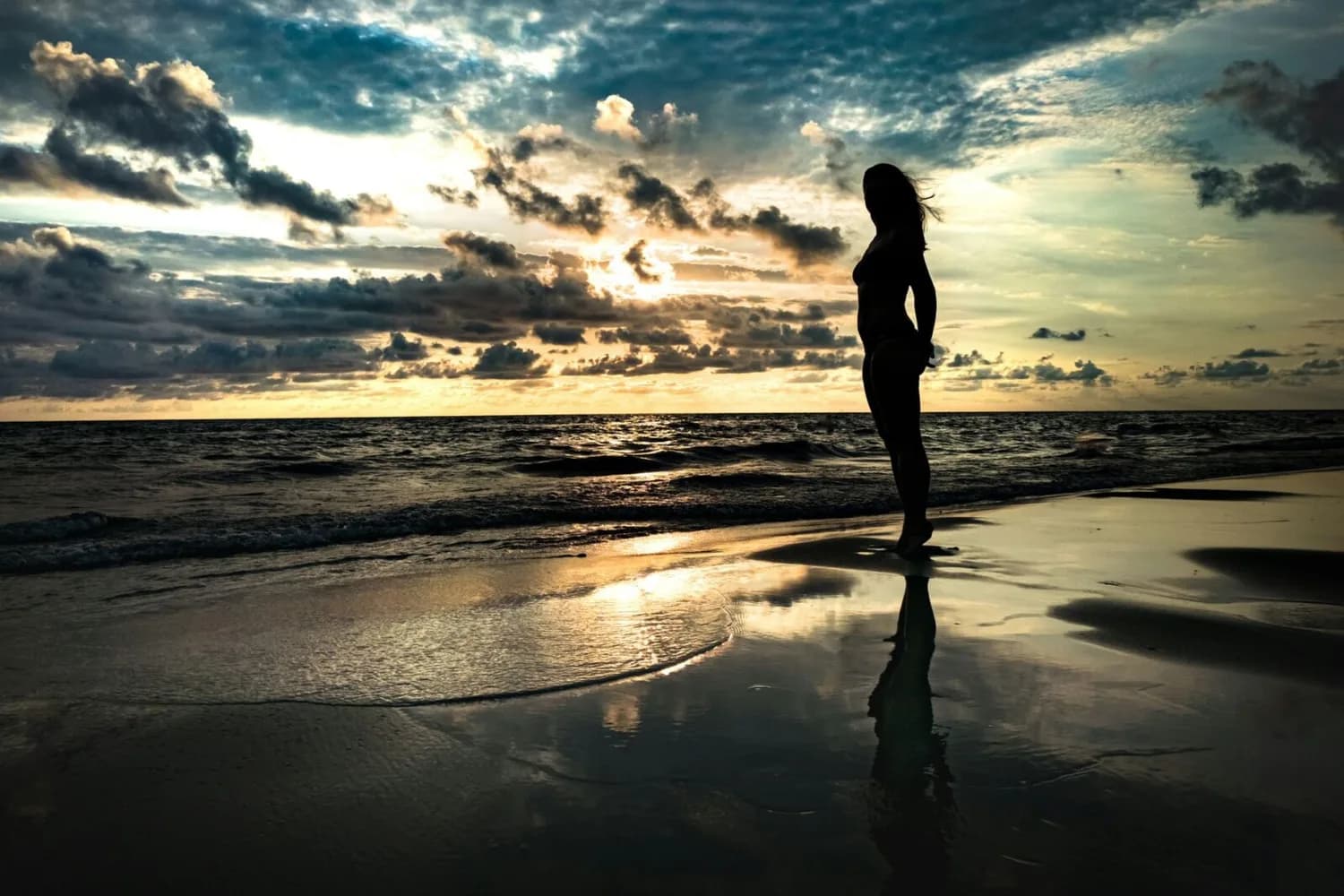 Silhouette einer Frau im Bikini am Strand bei Sonnenuntergang mit reflektierendem nassen Sand und dramatischen Wolken über dem Meer