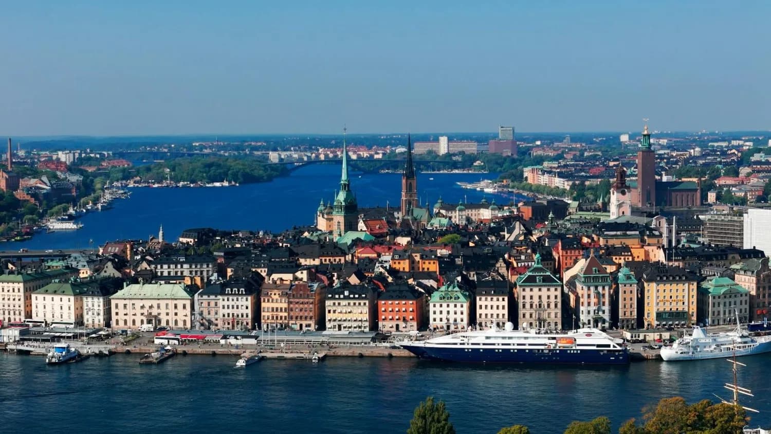 Panoramablick auf Stockholms Altstadt Gamla Stan mit bunten Häusern am Wasser, Kai mit Yachten und Kirchen- und Rathaus-Türmen im Hintergrund