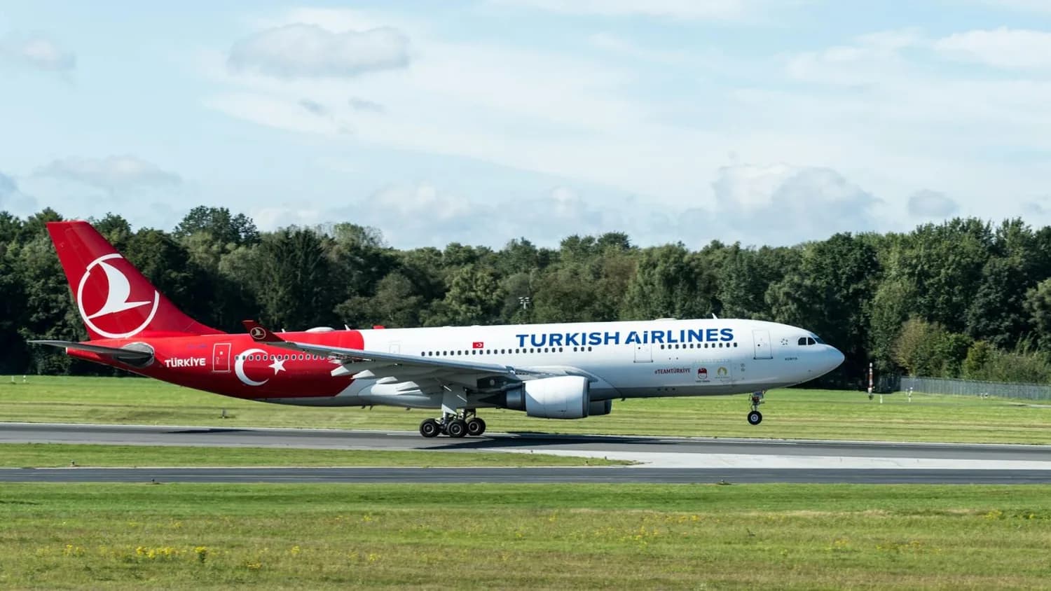 Turkish Airlines Passagierflugzeug mit rotem Heck und türkischem Halbmond landet auf Startbahn vor Grünfläche und Baumreihe