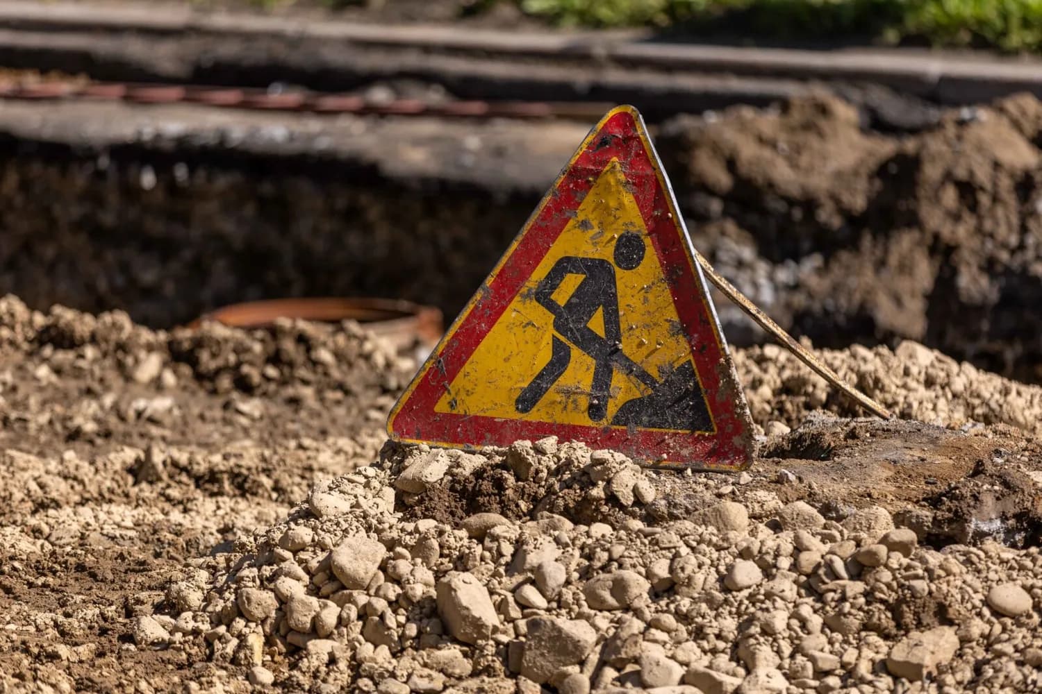 Warnschild Baustelle mit arbeitender Person auf Erdhaufen vor Graben und losem Schutt