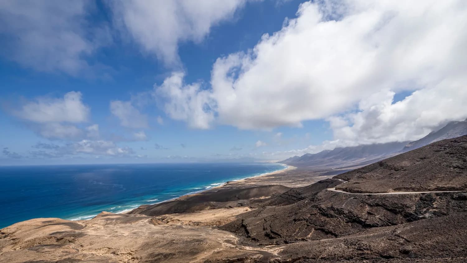 Luftaufnahme der vulkanischen Küste von Fuerteventura mit schwarzer Lavafelslandschaft, weitem Sandstrand und türkisblauem Atlantik unter bewölktem Himmel