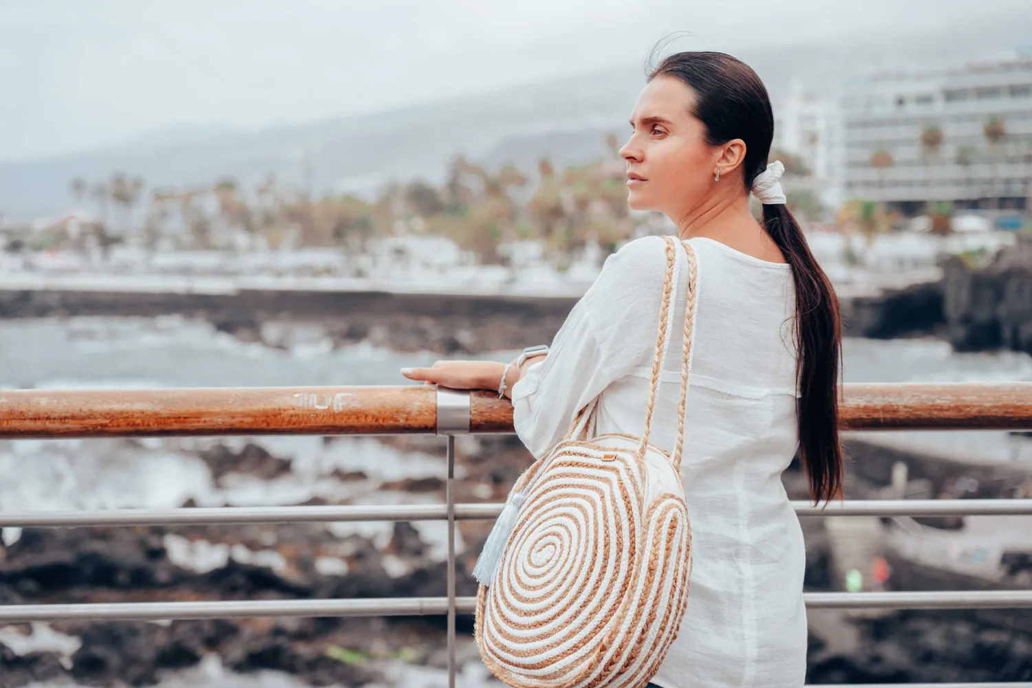 Frau in weißer Bluse mit geflochtener Strandtasche am Geländer mit Meerblick an der Küste von Puerto Cruz, Teneriffa