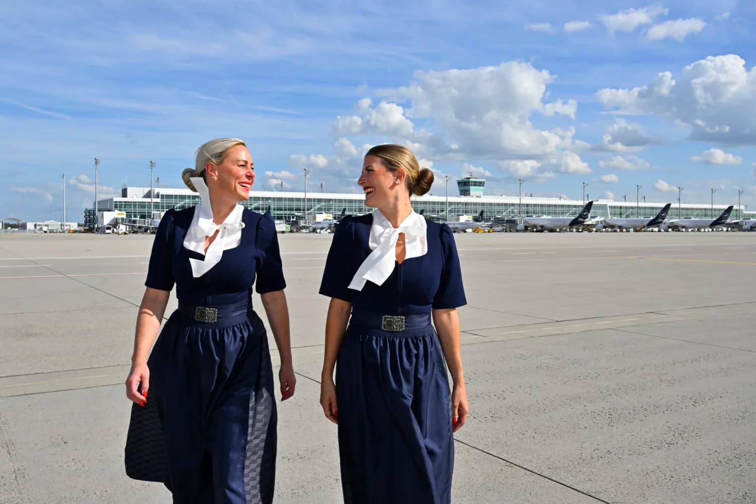Zwei lächelnde Lufthansa-Flugbegleiterinnen in traditioneller Tracht auf dem Rollfeld vor Terminal und geparkten Flugzeugen.