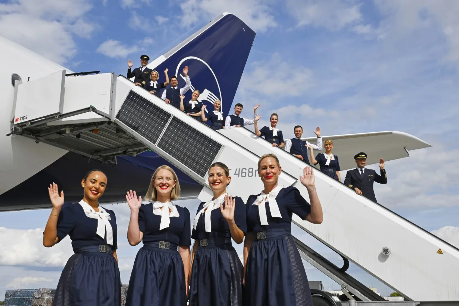 Lufthansa Kabinen- und Cockpitcrew in Trachtenuniformen posiert winkend an Flugzeugtreppe beim Oktoberfest-Motivshooting