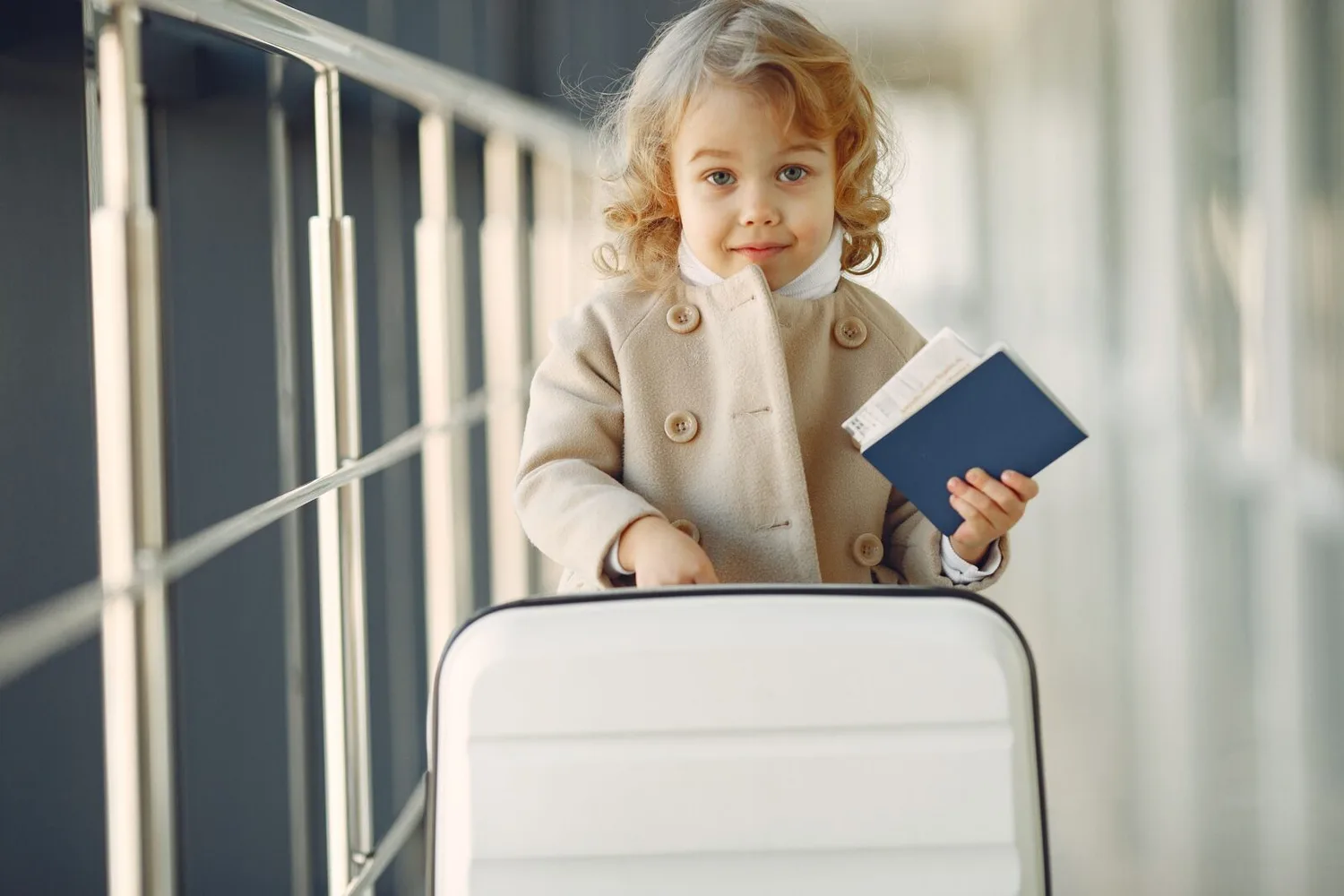 Kleines Kind im beigen Mantel am Flughafen mit Reisepass, Bordkarte und weißem Koffer
