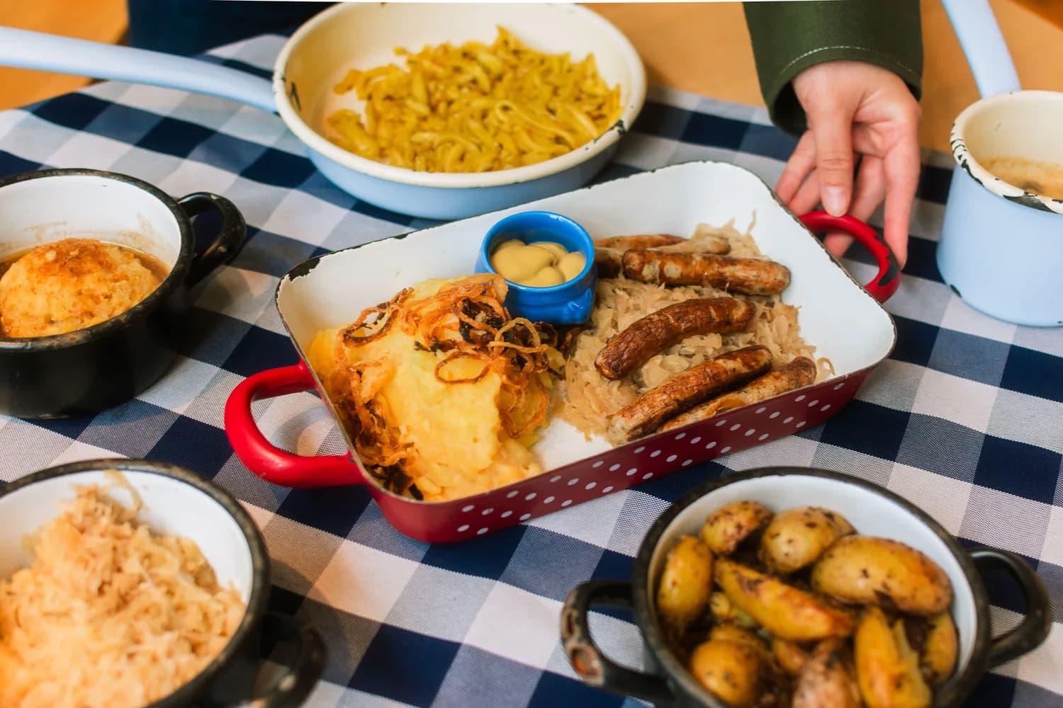 Bayerisches Schmankerl mit gebratenen Würstchen, Sauerkraut, Bratkartoffeln und Senf in Emaille-Blechschale auf kariertem Tischdecken-Arrangement