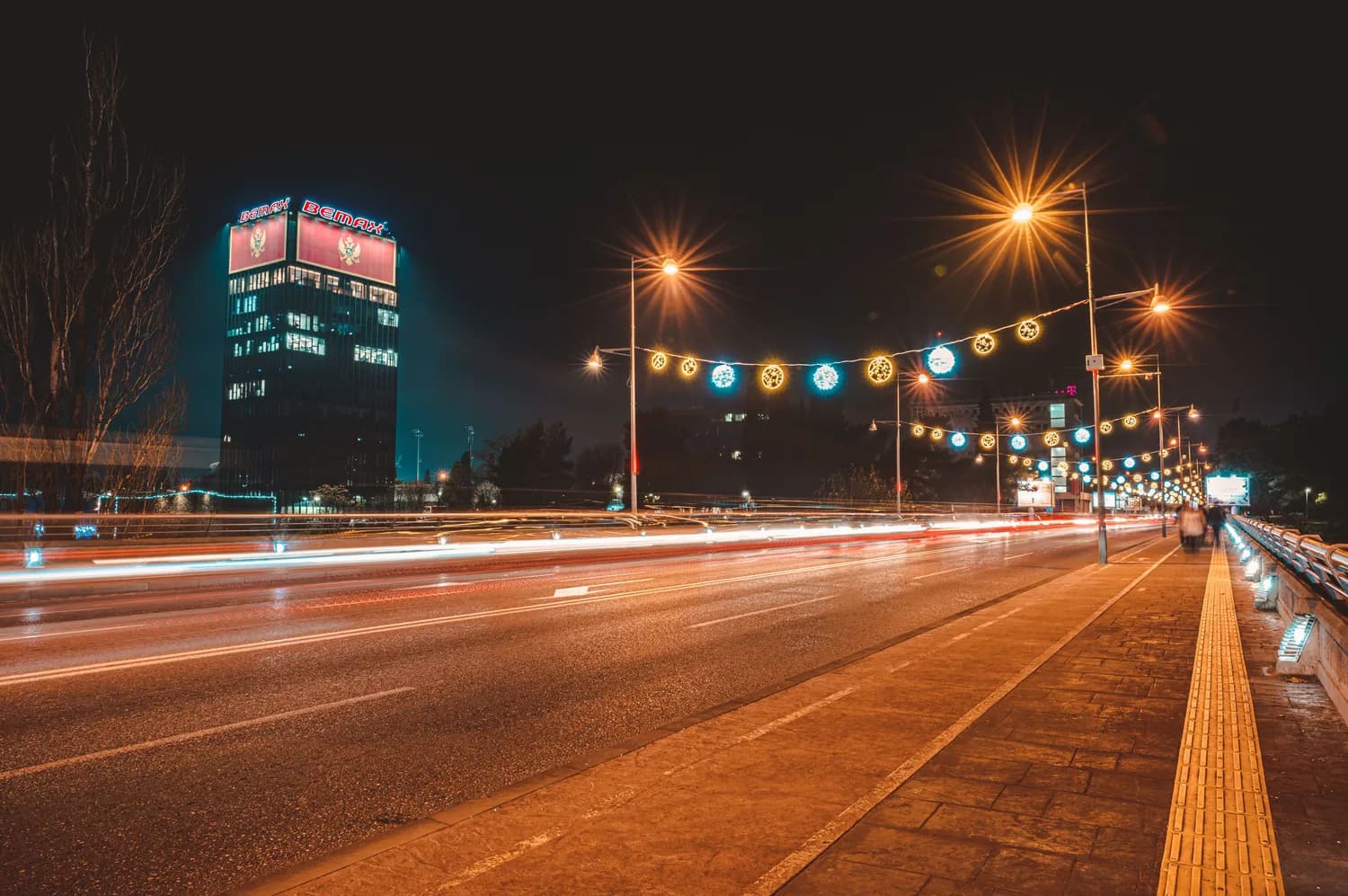 Nächtliche Straßenansicht von Podgorica Montenegro mit beleuchteter Hochhausfassade, festlichen Straßenlichtern und Lichtspuren vorbeifahrender Autos