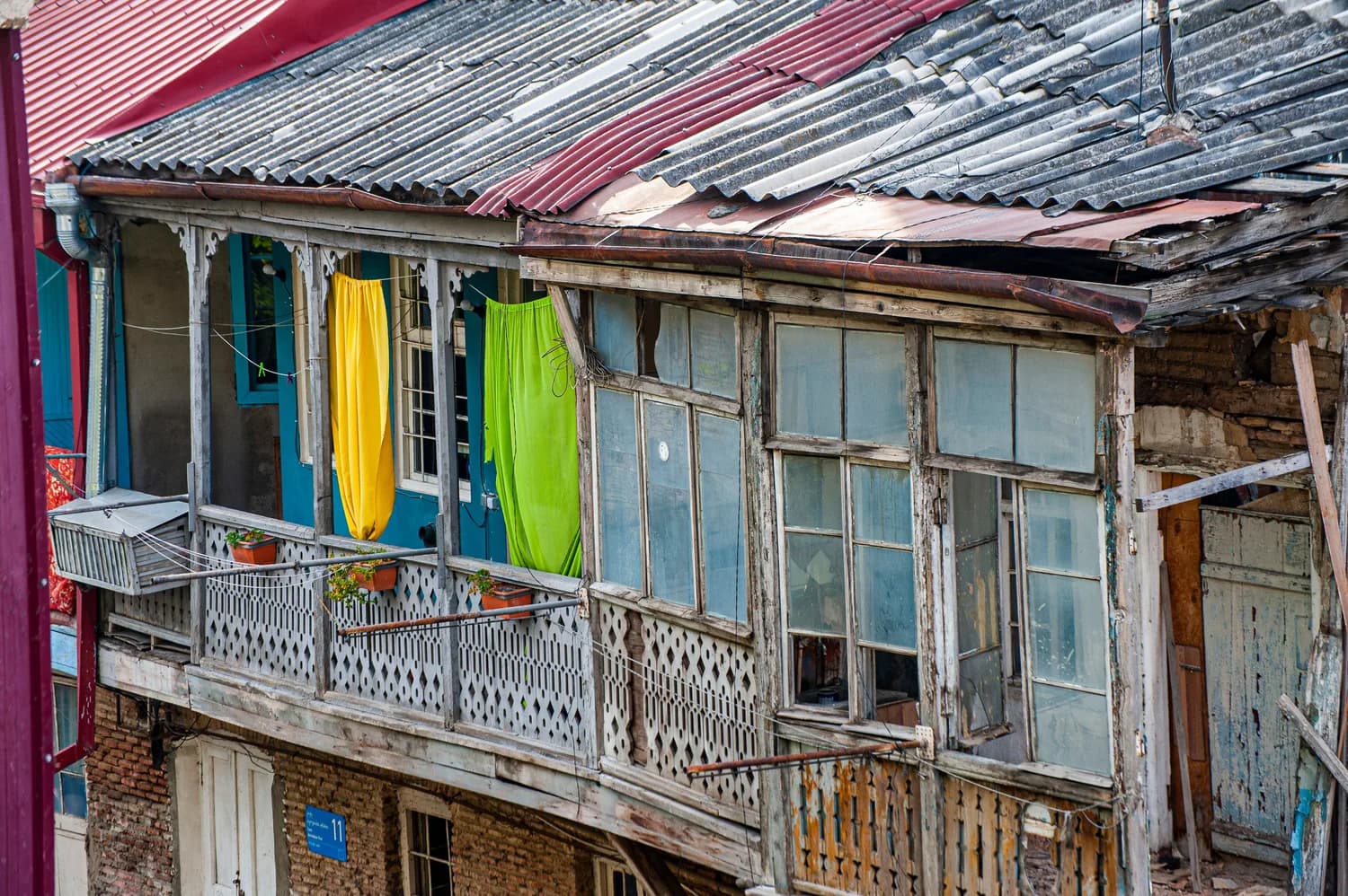 Rustikaler Holzbalkon in der Altstadt von Tiflis, Georgien mit bunten Vorhängen, verwitterten Fenstern und Pflanzenkübeln