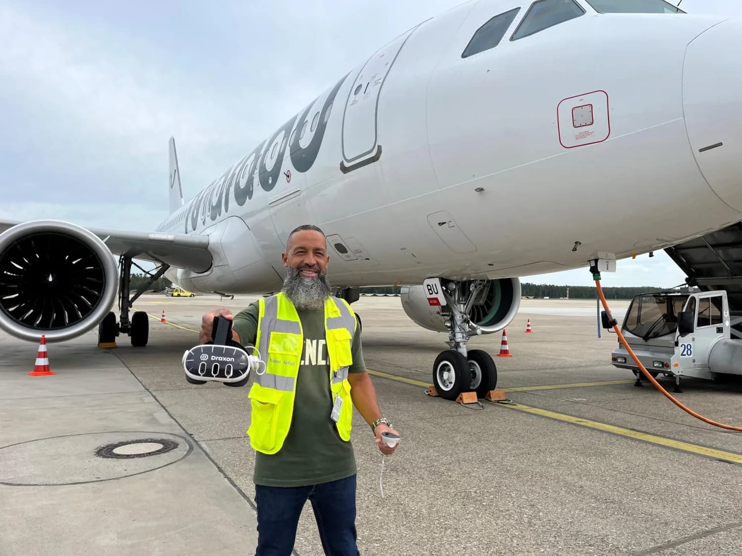 Ramp-Services-Trainer mit Warnweste vor passagierflugzeug hält Draxton Headset und Brille sichtbar.