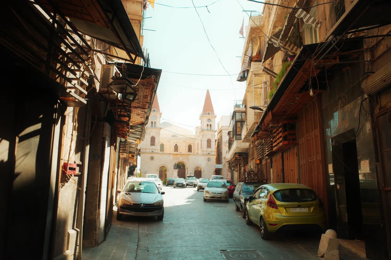 Engelse Straße in Aleppo mit geparkten Autos, engen Altstadtgassen und Kirche mit zwei Türmen im Hintergrund