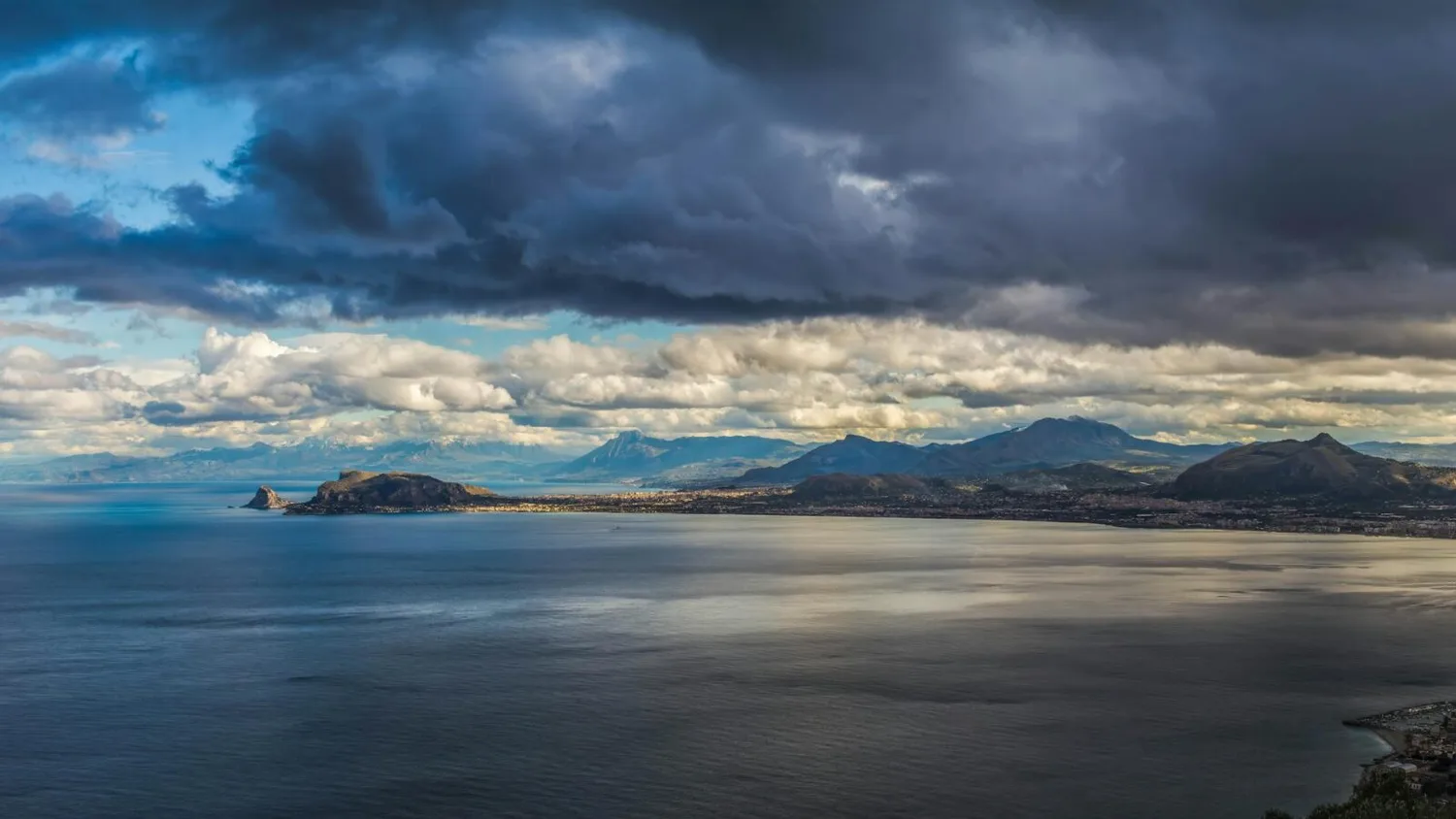 Küste bei Palermo auf Sizilien mit Strand, ruhigem Meer, vorgelagerten Felsen und dramatischen Wolken über Bergen und Küstenstadt