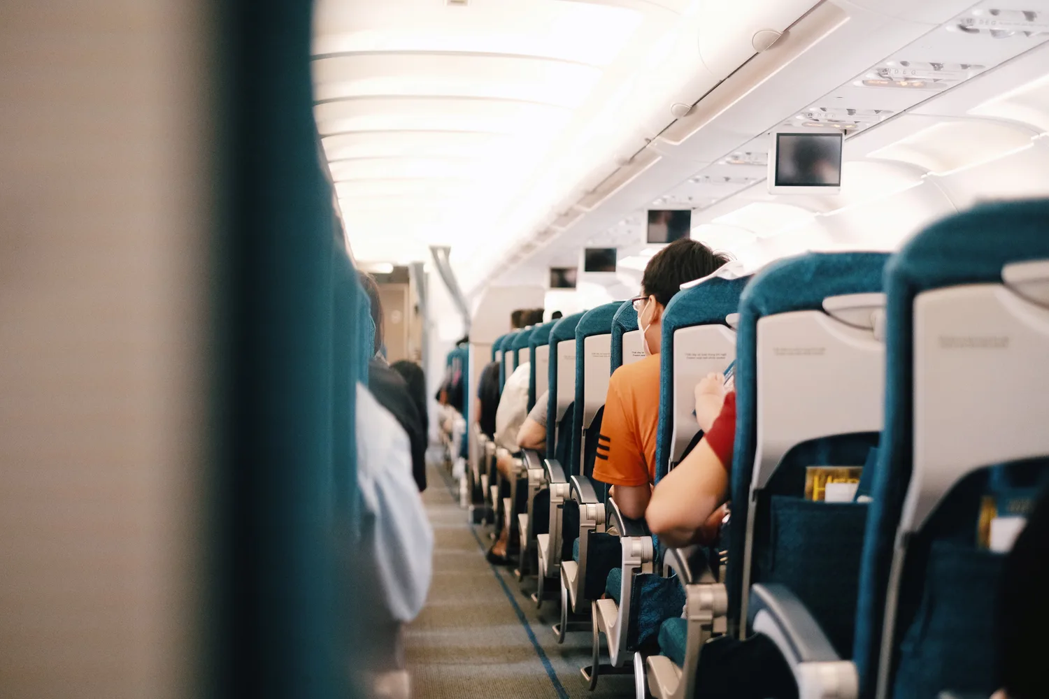 Innenansicht Flugzeugkabine mit sitzenden Passagieren auf Urlaubsziel Mallorca, schmale Economy-Reihe, Bordunterhaltung und Gepäckfächer sichtbar