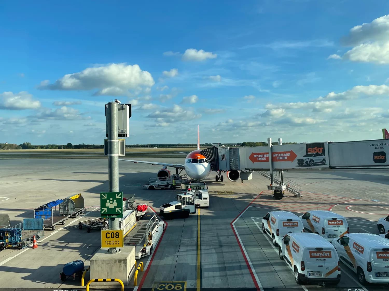 EasyJet Passagierflugzeug am Gate mit Fluggastbrücke und Bodenfahrzeugen am Flughafen Berlin-Schönefeld, Servicefahrzeuge mit EasyJet-Logo und Gepäckwagen auf dem Vorfeld