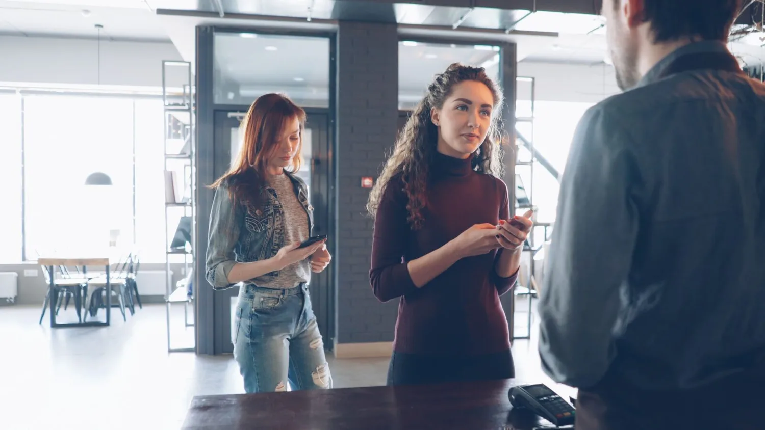 Zwei junge Frauen bezahlen mit Smartphone kontaktlos an einer Kasse im Café, Wartende Person im Hintergrund