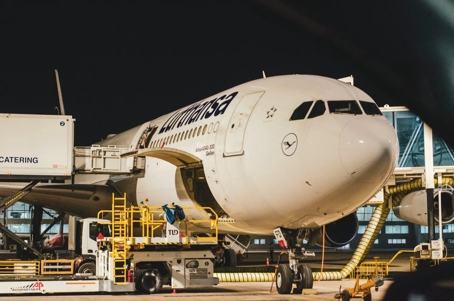 Lufthansa Airbus beim Boarding und Catering am Terminal, Flugzeugnase und Bodenservice mit Treppenwagen und Versorgungsleitungen bei Nacht