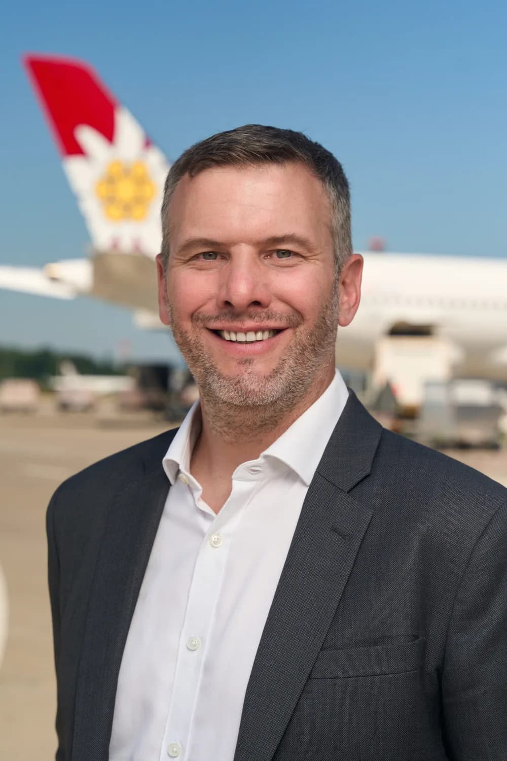 Lächelnder Mann im Anzug auf Flugplatz mit Flugzeugheck und Markenlogo im Hintergrund, Porträt von Edi Wolfensberger