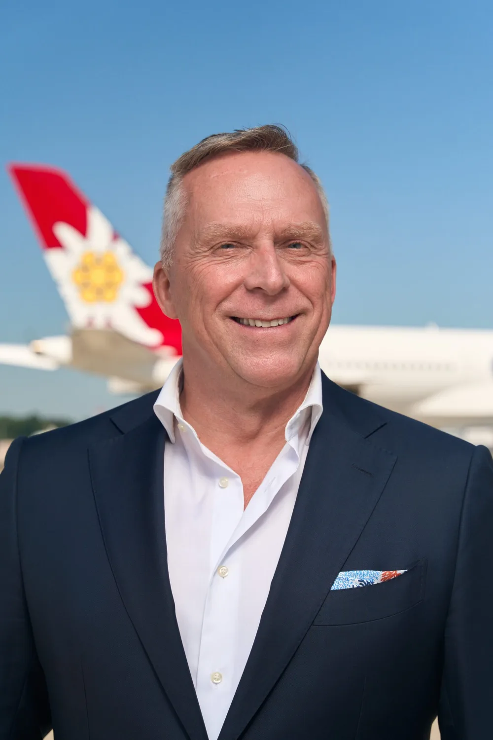 Porträt eines lächelnden Mannes im Anzug vor einem Flugzeug mit rotem Heck und Alpenblumen-Logo am Himmel