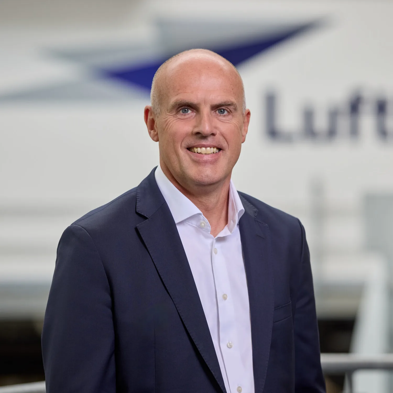 Mann im Anzug lächelt vor unscharfem Lufthansa-Logo, Business-Porträt in heller Büroumgebung