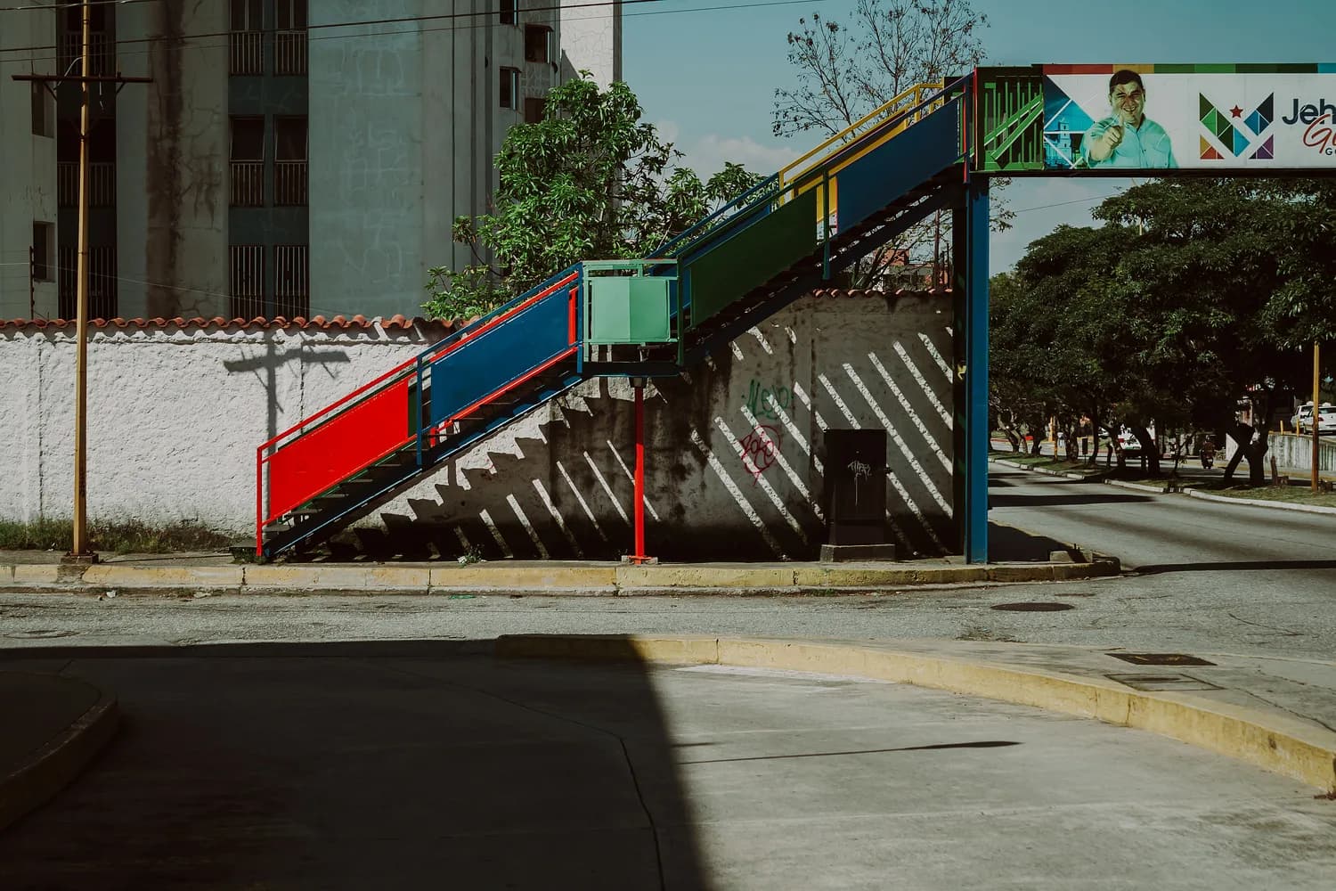 Bunte Fußgängerbrücke in Mérida, Venezuela mit roten, blauen und grünen Treppenabschnitten über einer Straße und Schattenmustern an einer weißen Mauer