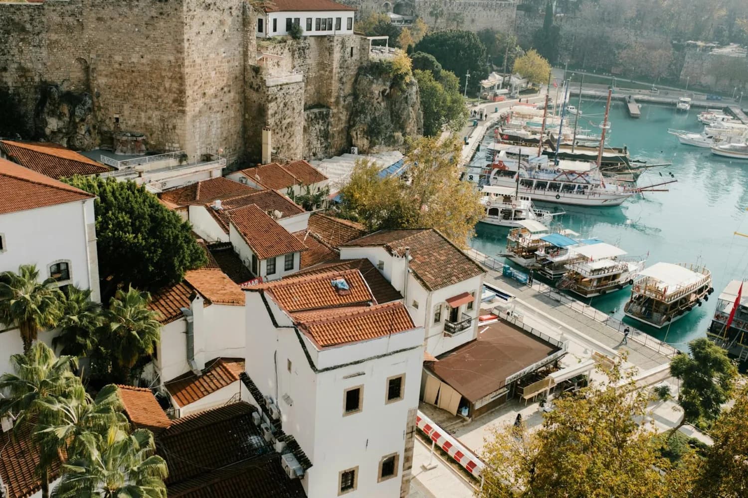 Hafen von Antalya mit weißen Häusern und roten Ziegeldächern, ankernden Ausflugsbooten und historischen Stadtmauern am türkisfarbenen Wasser