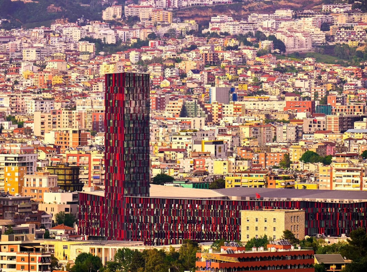 Panoramablick auf dicht bebautes Stadtviertel von Tirana, Albanien, mit markantem rotschwarzem Hochhaus und bunter Wohnbebauung am Hang