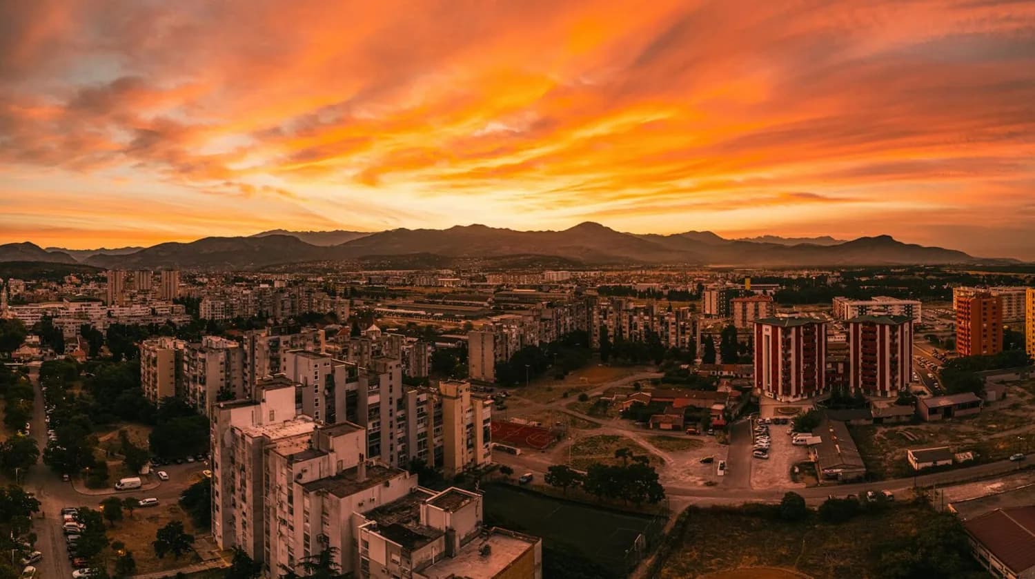 Panorama von Podgorica mit Wohnblöcken im Vordergrund, Stadtlandschaft und Bergen im Hintergrund bei dramatischem orangefarbenem Sonnenuntergang