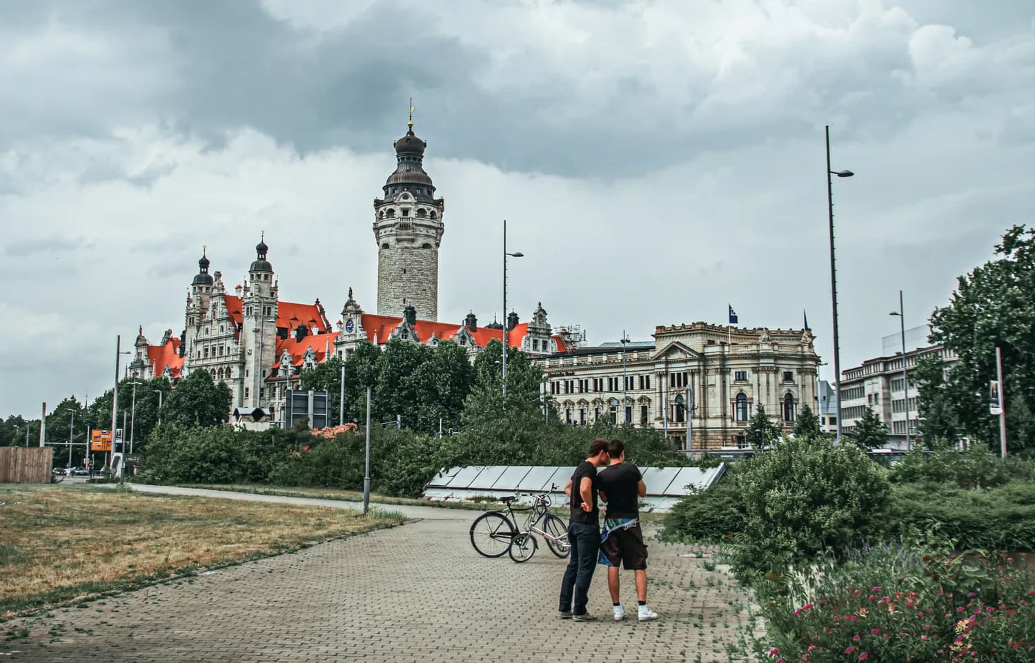 Panoramablick auf das historische Neues Rathaus Leipzig mit markantem Turm und roten Dächern, Männer mit Fahrrädern im Vordergrund, städtische Grünanlagen und bewölkter Himmel