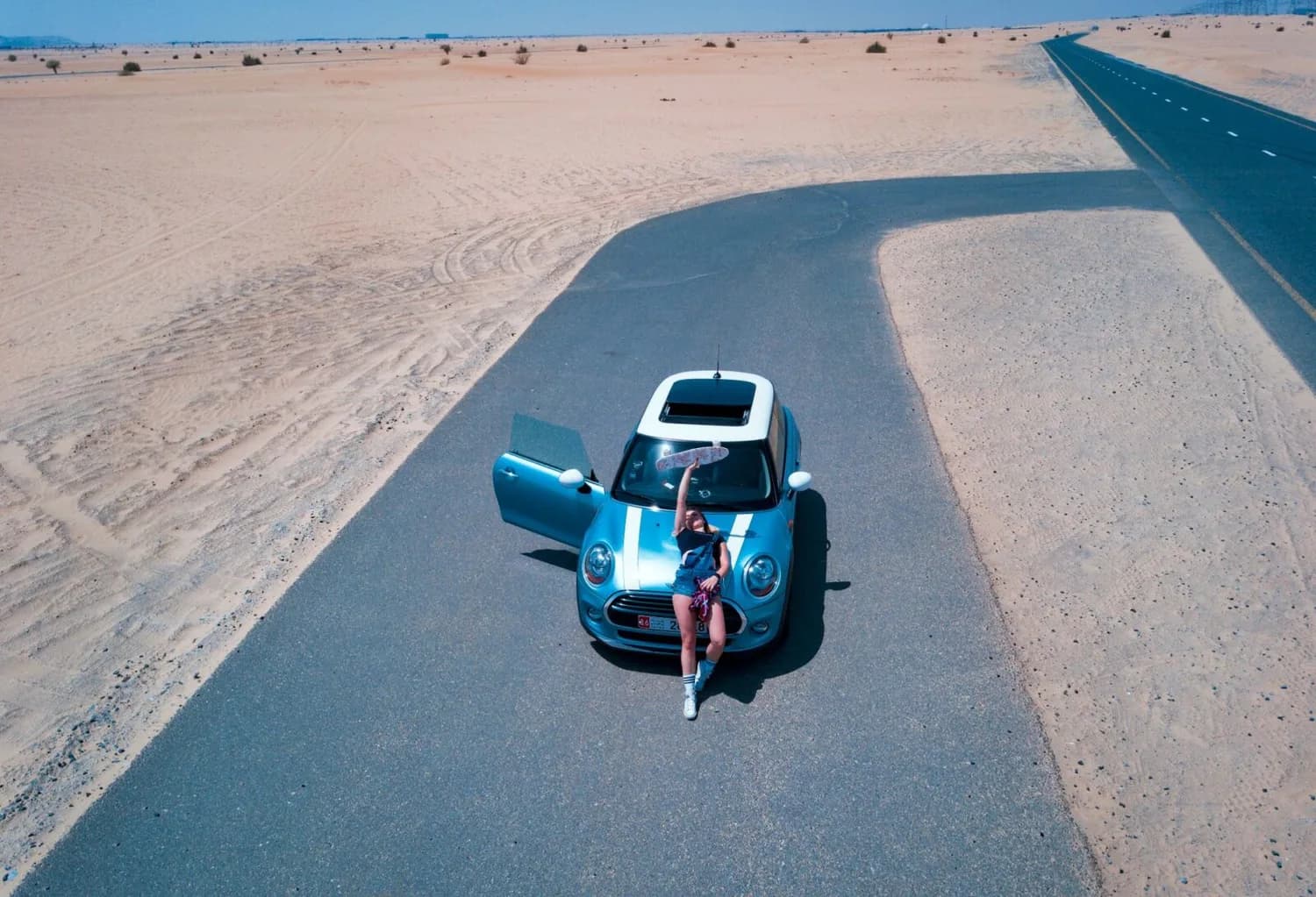 Frau lehnt an offener tür eines hellblauen Mini Cooper auf Wüstenstraße in Dubai mit Skateboard in der Hand und weitem Sandmeer im Hintergrund