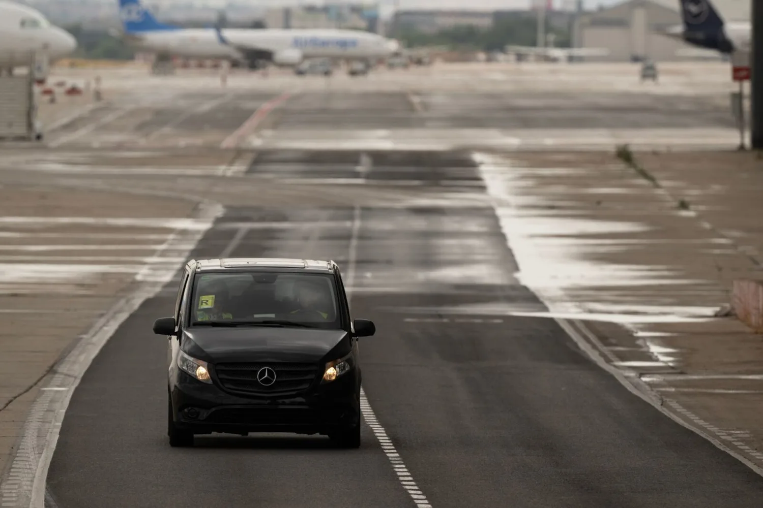 Schwarzer Mercedes Transporter fährt auf Rollweg am Flughafen, Flugzeuge und Startbahn im Hintergrund