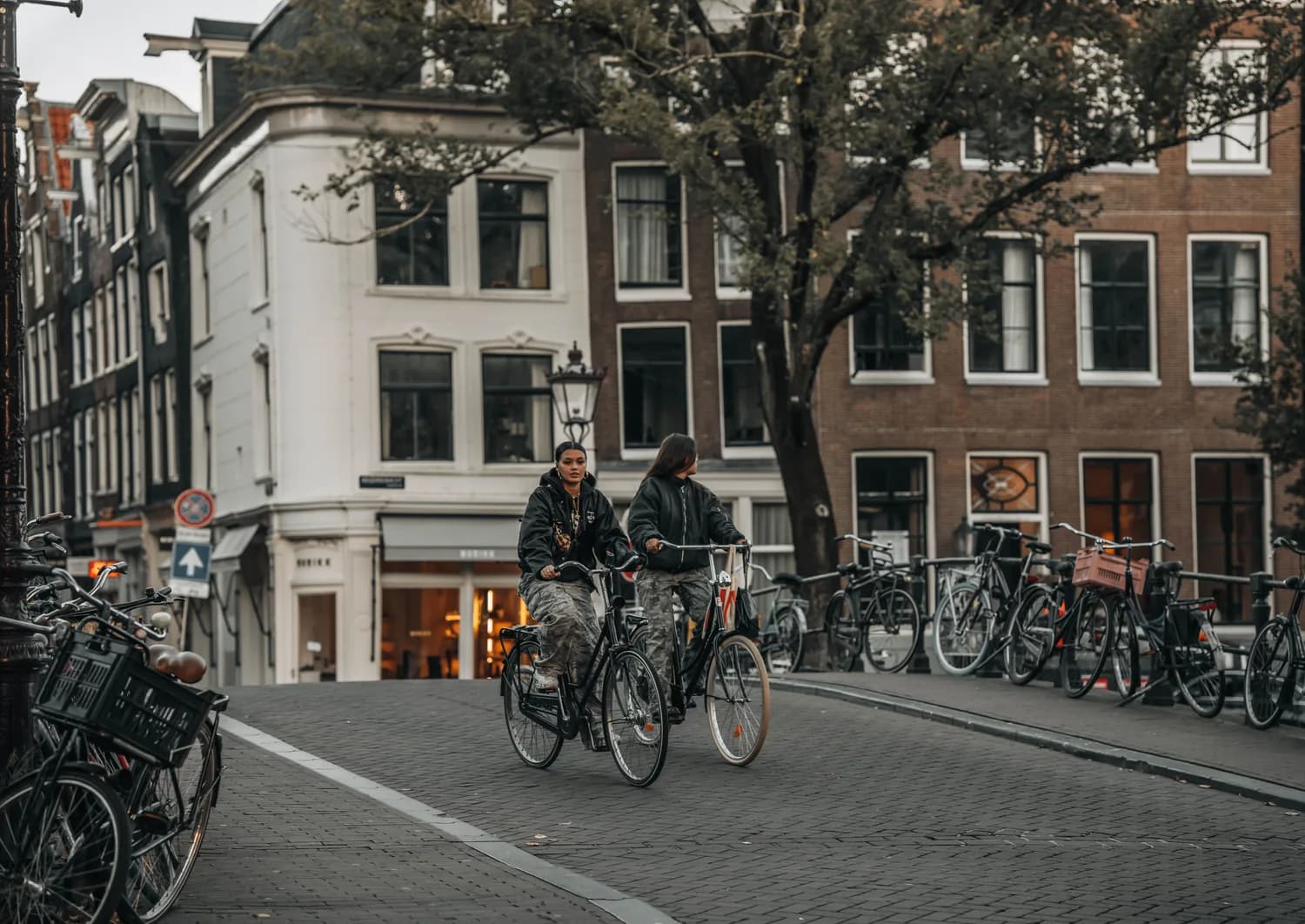 Zwei Fahrradfahrer auf einer Brücke über einen Amsterdamer Kanal vor historischen Giebelhäusern und abgestellten Fahrrädern