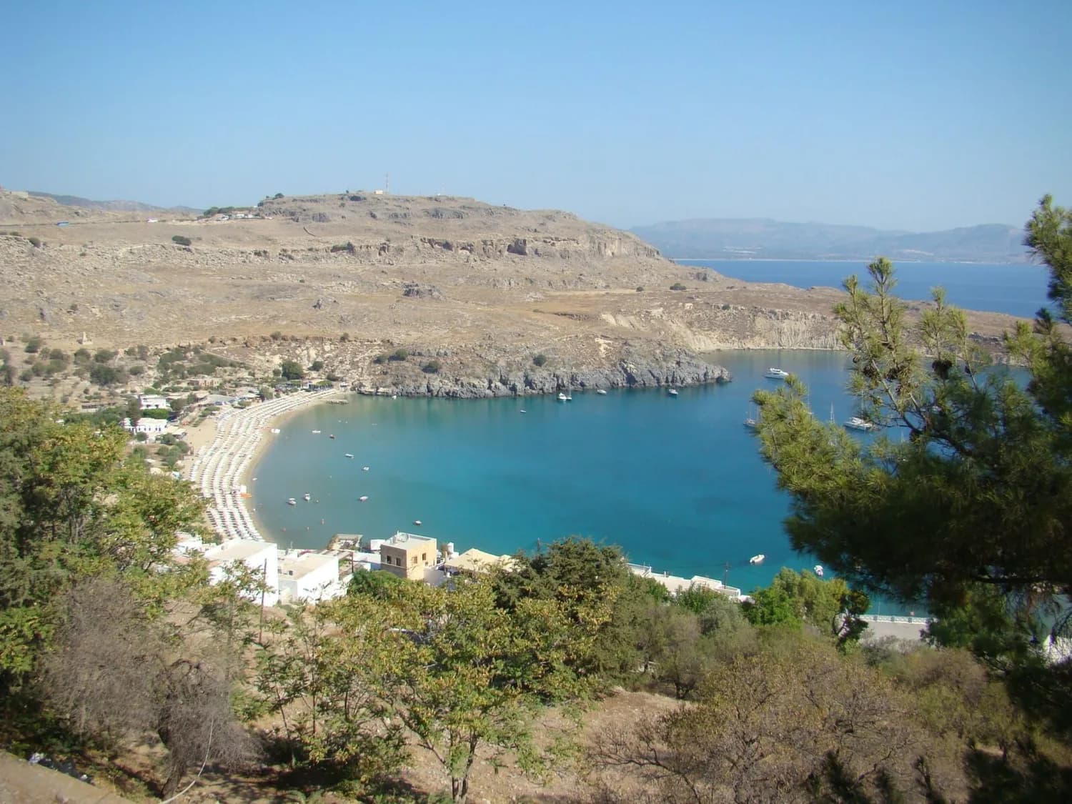 Bucht von Lindos mit türkisblauem Meer, Sandstrand mit Liegen, Klippen und Booten vor felsiger Küste in Griechenland