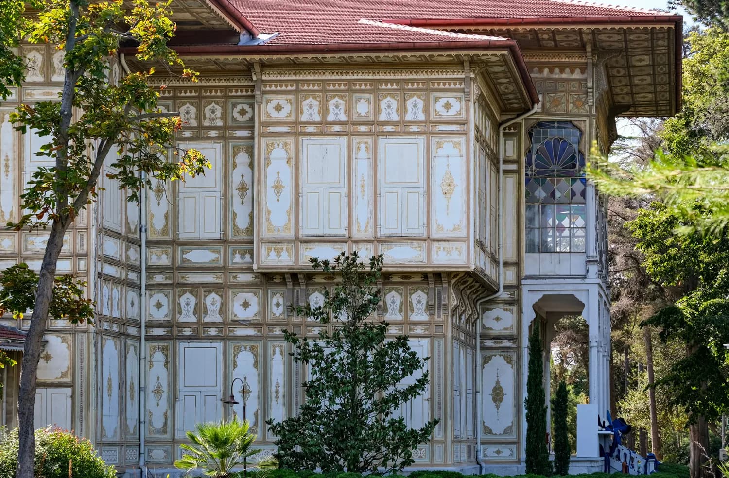 Verzierte Fassade eines historischen Holzhauses in Okurcalar mit filigranen Stuckornamenten, Erkerfenstern und bemalten Holzpaneelen, umgeben von Bäumen und Gartenpflanzen