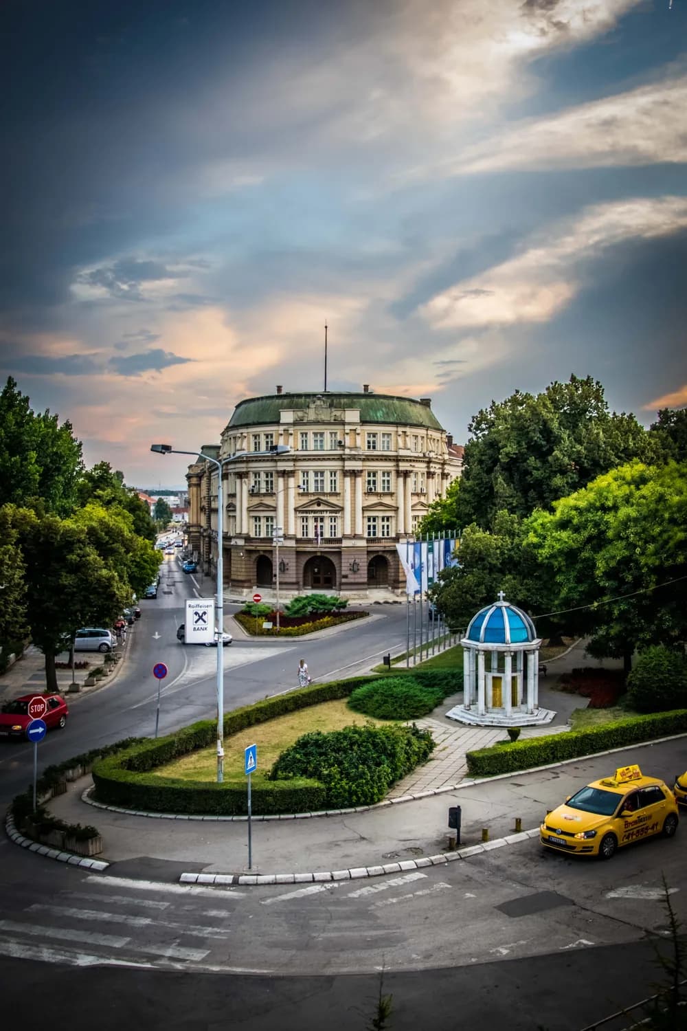 Historisches Rundbau-Gebäude in Niš, Serbien, Stadtplatz mit Pavillon, Bäumen und gelbem Taxi bei stimmungsvoller Abenddämmerung
