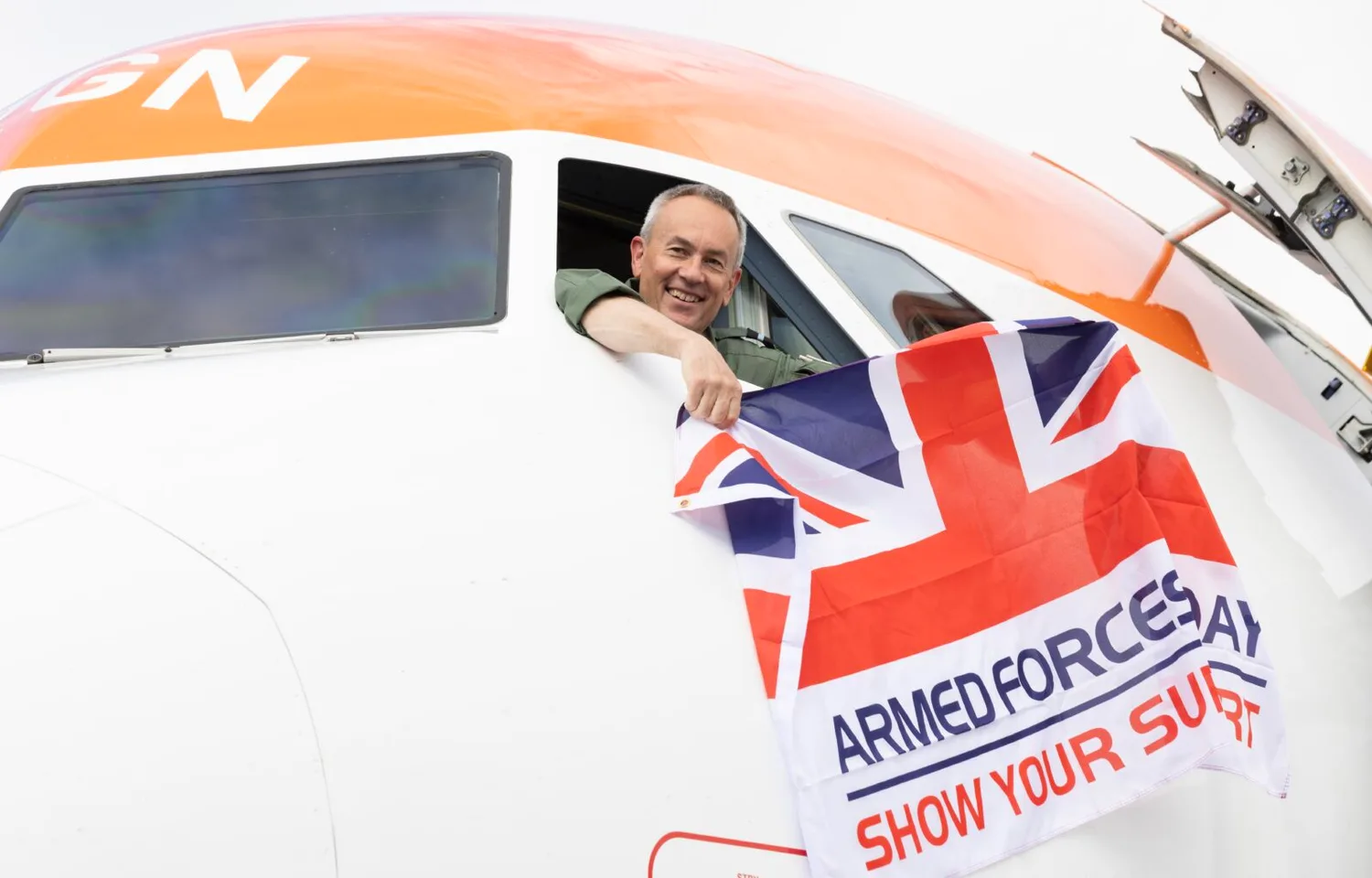 Pilot, lächelnd am Cockpitfenster eines Passagierflugzeugs, hält Union-Jack-Flagge mit Aufschrift Armed Forces Day - Show Your Support
