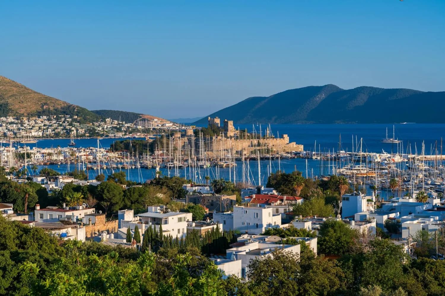 Panoramablick auf Bodrum Hafen mit zahlreichen Segelbooten, Yachten, weiß getünchten Häusern und der historischen Burg am Meer vor blauem Mittelmeer und Hügeln