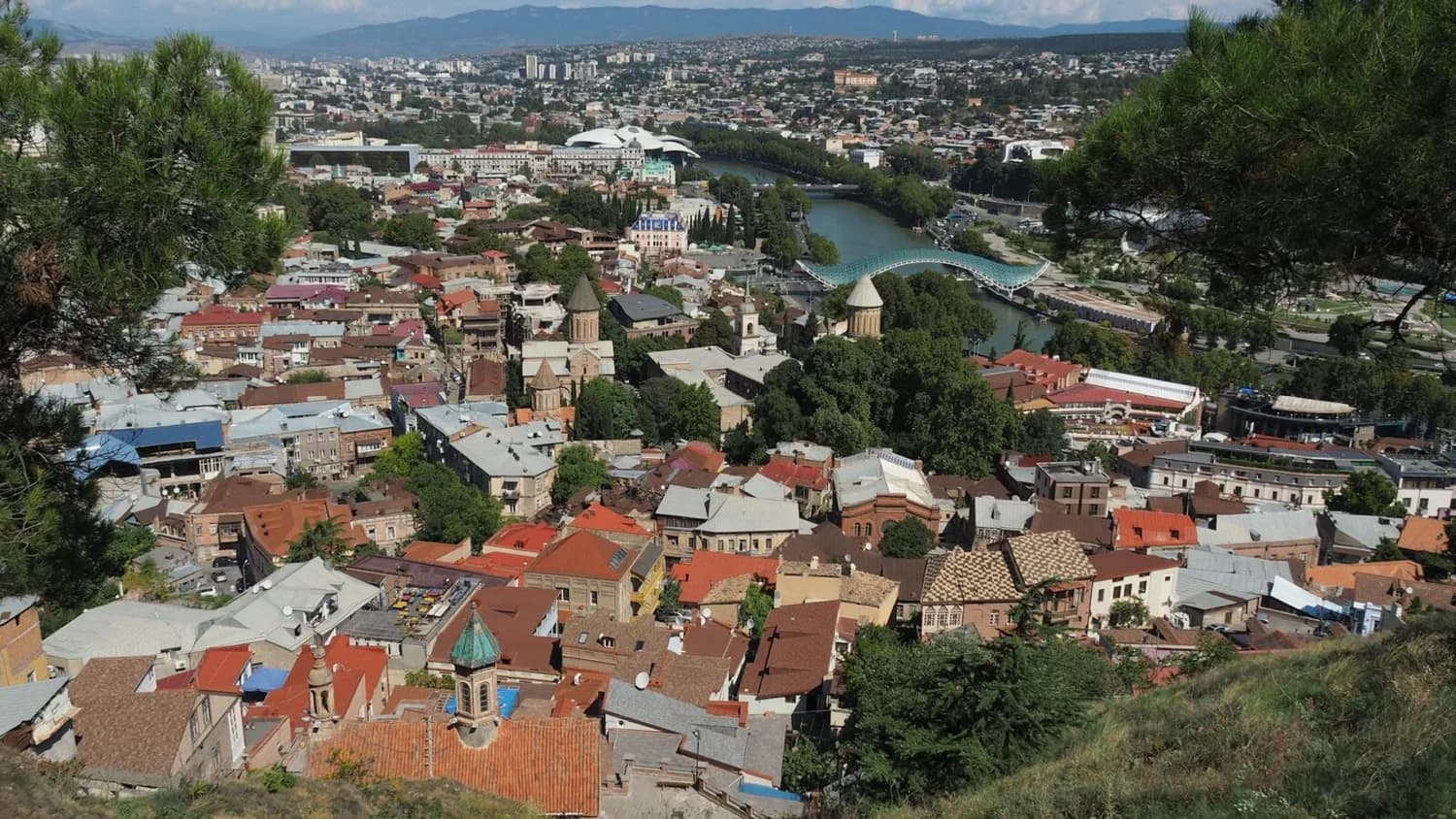 Panoramablick auf die Altstadt von Tiflis/Tbilisi mit bunten Dächern, orthodoxen Kirchen, Kura-Fluss und moderner Fußgängerbrücke, umgeben von Hügeln und Stadtlandschaft