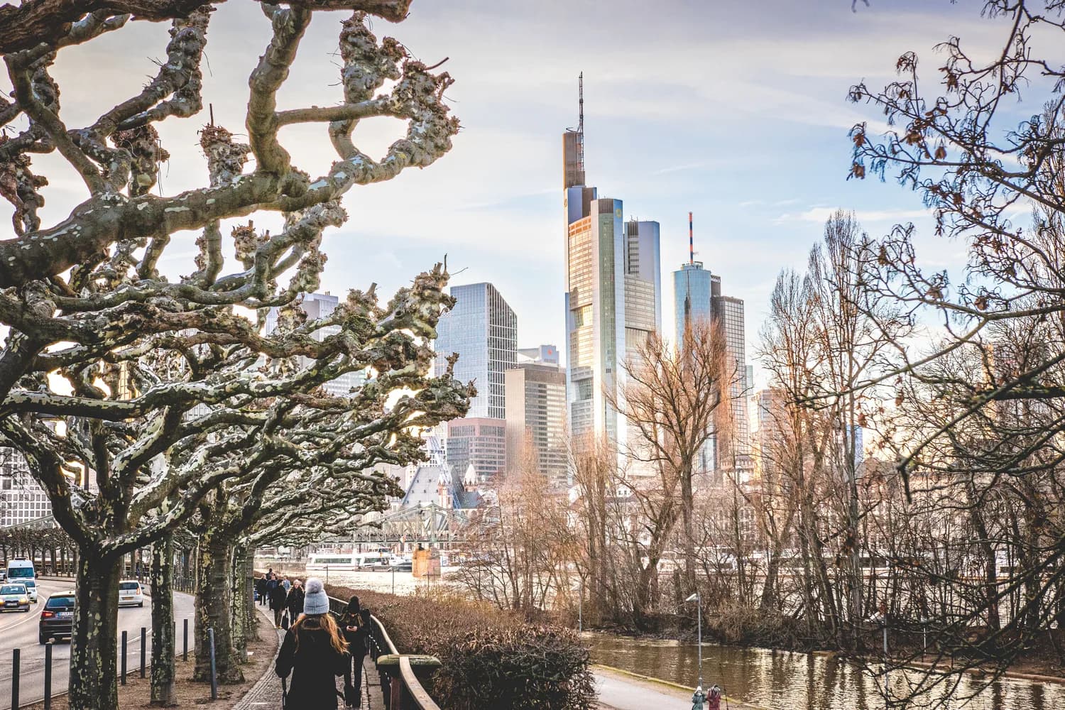Menschen spazieren am Winterufer des Mains mit Blick auf Frankfurts Skyline und Hochhäuser, kahle Platane-Bäume im Vordergrund