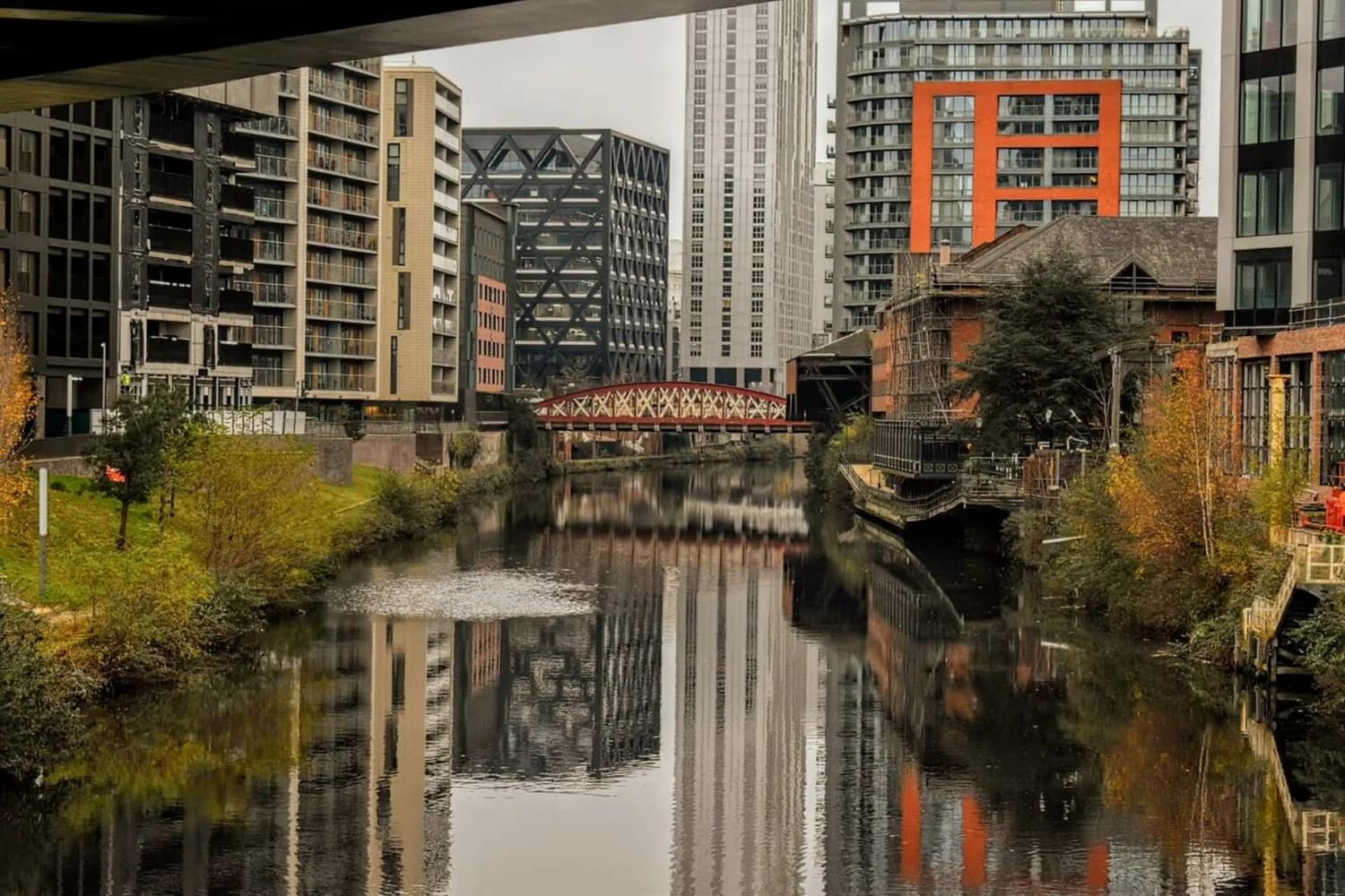 Blick auf den Manchester-Fluss Irwell mit Uferpromenade, modernen Wohn- und Bürogebäuden, roter Fachwerkbrücke und Spiegelung im Wasser