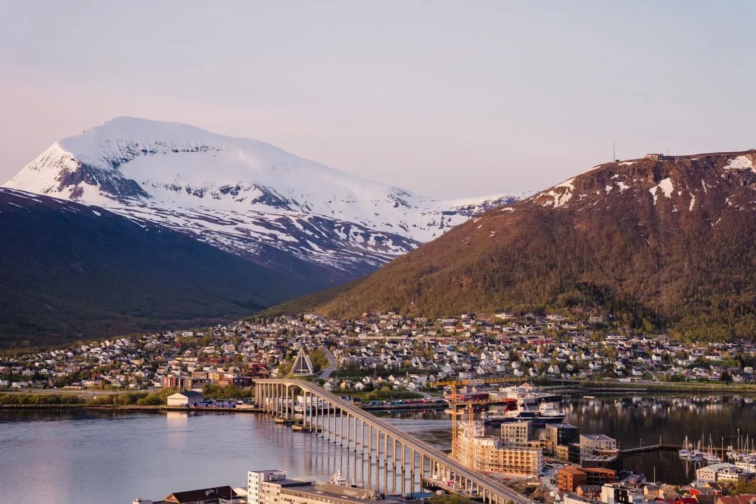 Blick auf Tromsø mit Tromsøbrua, Hafen, Wohnviertel und schneebedeckten Bergen im Abendlicht