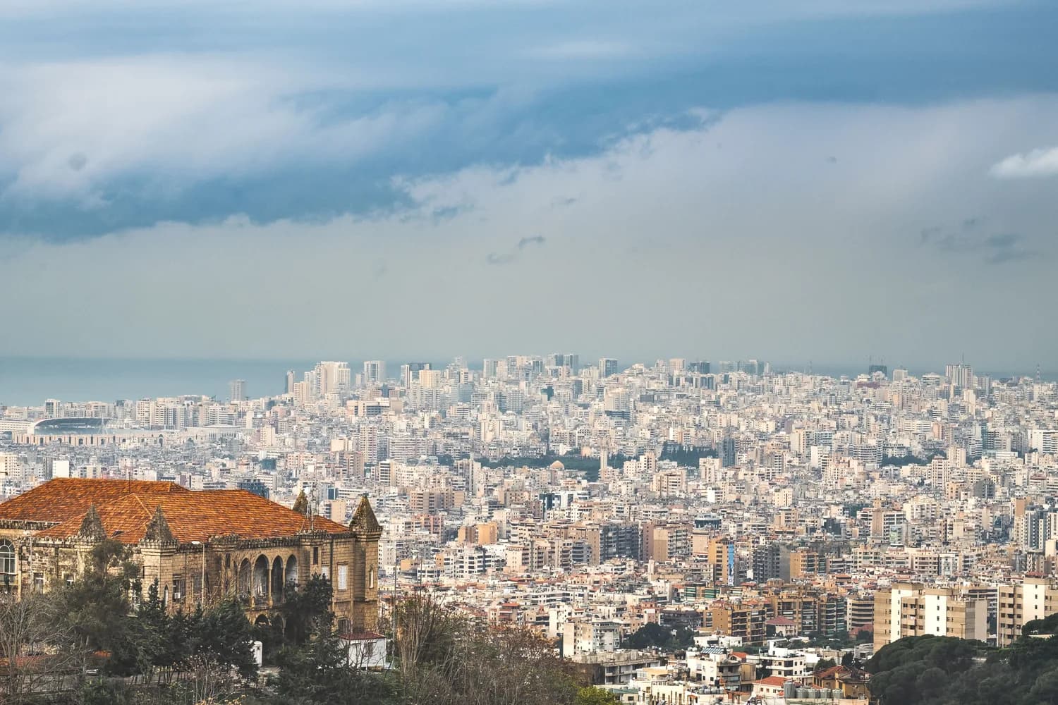 Panoramablick auf die dichte Skyline von Beirut mit vielen Wohn- und Hochhäusern, historische Villa mit roten Dach im Vordergrund, Mittelmeer im Hintergrund unter bewölktem Himmel