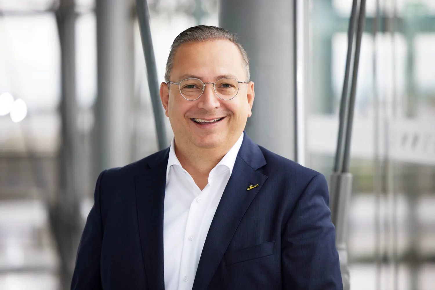 Mann im dunkelblauen Anzug und weißem Hemd lächelt vor modernem Glasgebäude, trägt runde Brille (Cenk Oezoeztuerk)